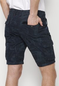 Sötét mintás cargo sort viselő férfi, akinek az egyik keze a hátsó zsebében van, egy egyszerű háttér előtt.