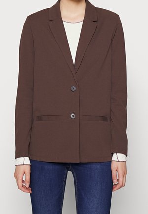 Kobieta ubrana w brązową marynarkę na dwa guziki, pod którą ma białą koszulę z długim rękawem i niebieskie dżinsy, stoi z rękami swobodnie opuszczonymi wzdłuż ciała.