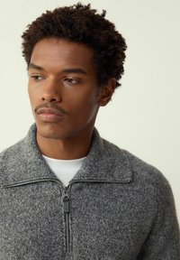 Lyhyttukkaisen kiharatukkaisen miehen yllään harmaa vetoketjullinen neulepusero valkoisen paidan päällä, katse vasemmalle, vaaleaa taustaa vasten.