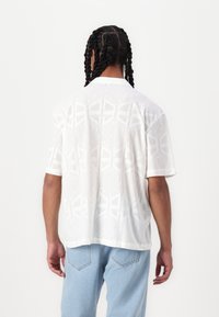 Chemise blanche à manches courtes avec un motif géométrique texturé et un tissu doux et léger. Le col présente un accent de design subtil.