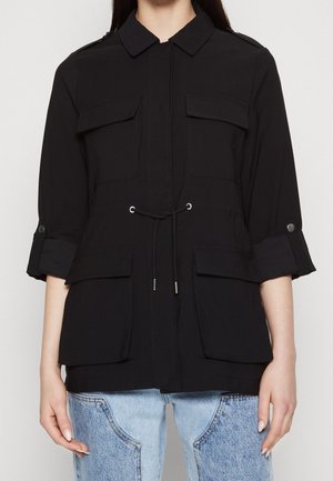 Vrouw die een zwarte jas met knopen draagt, met klepzakken op de borst en heupen, opgerolde driekwart mouwen en een taille met trekkoord, gecombineerd met een blauwe spijkerbroek.