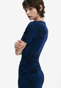 Mörkblå klänning med figurnära silhuett, korta ärmar och rynkade detaljer vid midjan. Tyget ser slätt ut med subtila svarta mönster.