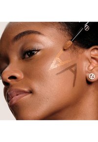 Gros plan sur une femme appliquant un correcteur liquide beige en forme de "V" sur sa joue près de l'oreille avec un applicateur de maquillage.