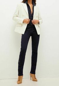 Valkoinen räätälöity bleiseri, tummat farkut hopeakoristeilla ja vaaleat korkokengät. Asusteessa on syvä V-pääntie bleiserin alla.