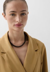 Hellbrauner Blazer mit geknöpften Revers, strukturiertem Stoff, kombiniert mit einer schlichten schwarzen Perlenkette. Glatte Haut und dezentes Make-up.