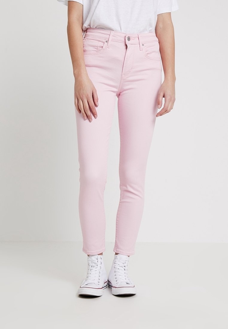 Femme portant un jean skinny rose clair taille haute et des baskets blanches montantes, debout devant un fond blanc uni.