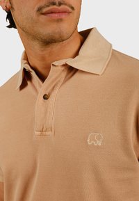 Beige polohemd van textiel met structuur, met een drie-knopenlijntje en een geborduurd olifantenlogo op de linkerkant van de borst.