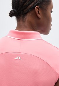 Άτομο με πλεξούδες που φοράει ροζ πουκάμισο με γιακά, με λευκό μικρό λογότυπο στο πάνω μέρος της πλάτης, γυρισμένο πλάτη στην κάμερα.