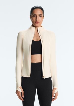 Donna con capelli scuri raccolti indietro, indossa una giacca beige chiaro con cerniera aperta, un reggiseno sportivo nero e leggings neri a vita alta.