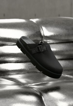 Zapato negro sin cordones con parte superior de cuero texturizado, con detalle de correa y hebilla, y una suela gruesa y resistente. Fondo metálico brillante.