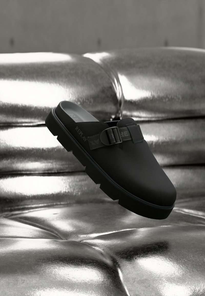 Μαύρο παπούτσι slip-on με υφή δερμάτινου επάνω μέρους, που διαθέτει λουράκι και πόρπη, και παχιά, ανθεκτική σόλα. Γυαλιστερό μεταλλικό φόντο.