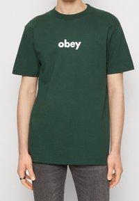 T-shirt en coton vert avec des manches courtes et un col rond ; présente le texte blanc "obey" imprimé sur le devant. Texture lisse, coupe décontractée.