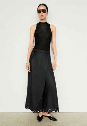 Donna con occhiali da sole, top nero senza maniche, ampia gonna nera a portafoglio con orlo intagliato, ballerine nere e braccialetti d’argento, in piedi al chiuso.