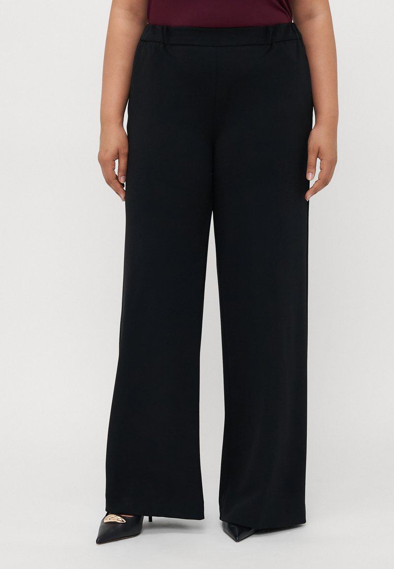 Mujer vestida con pantalones de pierna ancha negros y tacones altos puntiagudos negros con detalle dorado, de pie frente a un fondo blanco liso.