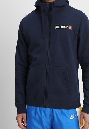 Granatowa bluza z kapturem i suwakiem, z napisem "JUST DO IT." na piersi, noszona z jasnoniebieskimi szortami Nike z białym logo.