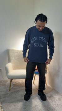 Man standing indoors looking down, wearing a navy blue "New York State" long-sleeve shirt, black pants, and black shoes near a white armchair.