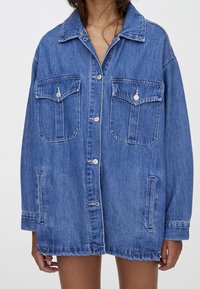 Mujer vestida con una chaqueta de mezclilla azul oversized, con botones en el frente y cuatro bolsillos, de pie frente a un fondo claro y liso.