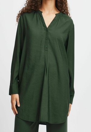 Vrouw draagt een donker groene tuniek met lange mouwen, een button-down V-hals en een geplooide halslijn, getoond van schouders tot halverwege de dijen.
