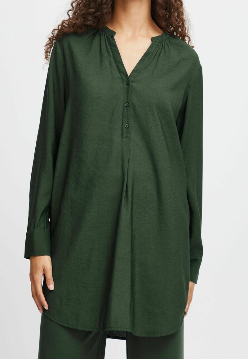 Femme portant une tunique à manches longues vert foncé avec un col en V boutonné et un col froncé, montrée des épaules jusqu'à mi-cuisse.