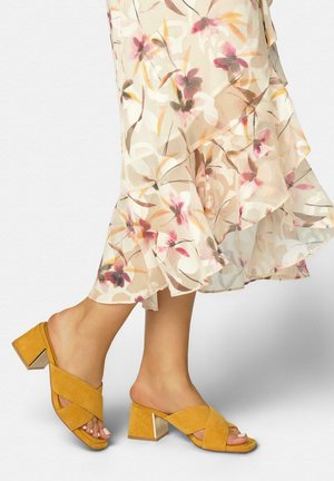 Mujer que lleva una falda con volantes florales y sandalias de tacón ancho en mostaza con tiras cruzadas en los pies descalzos.