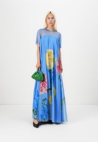 Light blue maxi dress with floral print, sheer overlay, and short sleeves. Green pleated handbag adds contrast. Black shoes complete the outfit.