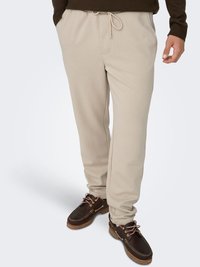 Beige sweatpants with an elastic waistband and drawstring detail. Smooth fabric, relaxed fit, paired with dark brown shoes and pink laces.