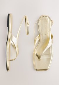 Sandali piatti oro metallizzato dal design elegante, con cinturini incrociati, punta quadrata e una sottile suola nera.