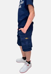 Pantaloni cargo blu navy con tasche laterali e un distintivo ricamato, abbinati a sneakers bianche con accenti rossi e calze nere.
