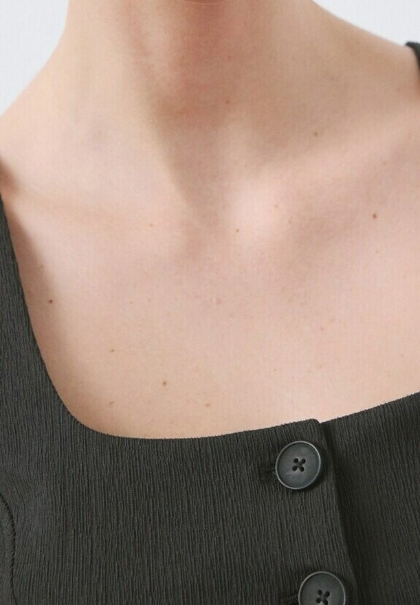 Gros plan sur le cou et le haut de la poitrine d'une personne portant un haut noir texturé avec de gros boutons noirs.