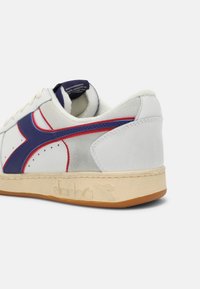 Les baskets Diadora présentent un cuir blanc et du daim gris, avec des accents rouges et bleus. La semelle en caoutchouc est beige avec un logo embossé sur le côté.