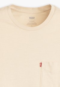 Beige Levi's t-shirt med rund halsudskæring, korte ærmer og brystlomme. Har et lille rødt logo mærke på lommen. Stoffet ser blødt ud.