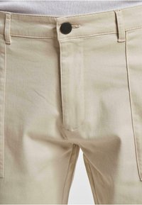 Beige kangashousut, joissa on sileä pinta, varustettu metallinappikiinnityksellä, etutaskuilla ja ommelluilla yksityiskohdilla saumoissa.