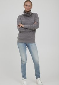 Šedá mikina s vysokým límcem a strukturovaným vzorem zig-zag, vyznačující se dvěma postranními knoflíky. Spárována s světle modrými džíny a bílými teniskami.
