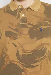 Brun och beige kamouflagemönstrad polo med en beige krage och en liten blå broderad logotyp på vänster bröst.