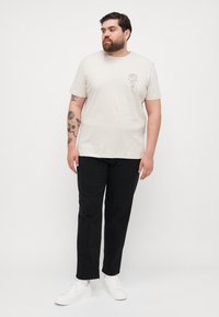 Beige cotton t-shirt with a floral outline graphic on the left chest, paired with black pants and white sneakers.