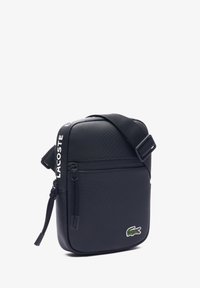 Bolso negro de bandolera hecho de nylon texturizado, con un bolsillo frontal con cremallera, correa ajustable y un logo de cocodrilo bordado en verde.