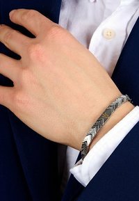 Metallarmband med ett geometriskt mönster och växlande texturer, silvrig finish, och en liten präglad emblem, bärs på en hand.