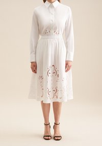 Hvid skjortekjole med lange ærmer, knaplukning foran, samlet talje og blonderlignende broderi i kanten. Kombineret med sorte hæle sandaler.