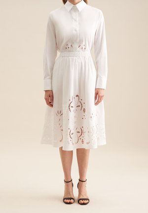 Witte jurk met lange mouwen, knopen aan de voorkant, geplooide taille en kantachtige borduursels aan de zoom. Gecombineerd met zwarte hoge hakken sandalen.