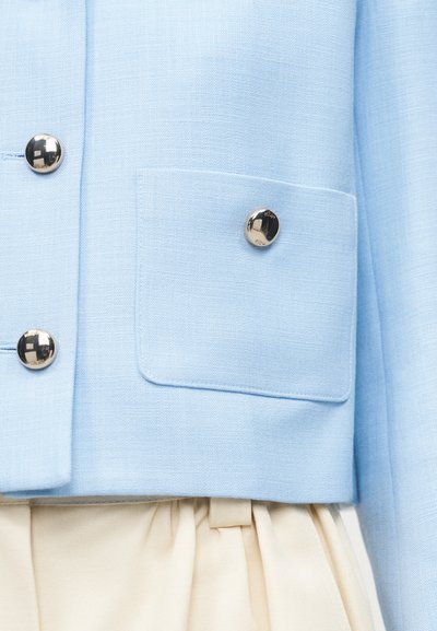 Ανοιχτό μπλε μπουφάν με ασημί στρογγυλά κουμπιά και μπροστινή τσέπη, συνδυασμένο με κρεμ πλισέ ύφασμα από κάτω.