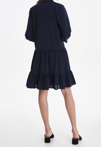 Abito blu navy con orlo a balze stratificate, maniche lunghe e colletto con bottoni. Realizzato in tessuto leggero con una texture liscia.