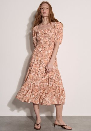 Kobieta o długich rudych włosach, ubrana w różową sukienkę midi w paisley z bufkami na rękawach i kwadratowym dekoltem, pozująca na tle jednolitej ściany.