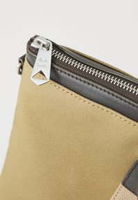 Tan stofpose med en mørkebrun læderdetalje, der har et metal lynlås træk med indgraveret branding. Bemærkelsesværdige tekstur- og syningsdetaljer.