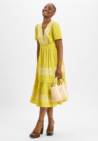 Yellow cotton dress with short sleeves, floral embroidery, tiered skirt, and a V-neck. Paired with brown platform sandals and a woven bag.