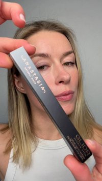 Femme blonde à la peau claire tenant une boîte de crayon à sourcils Anastasia Beverly Hills Glider près de son visage sur un fond gris uni.