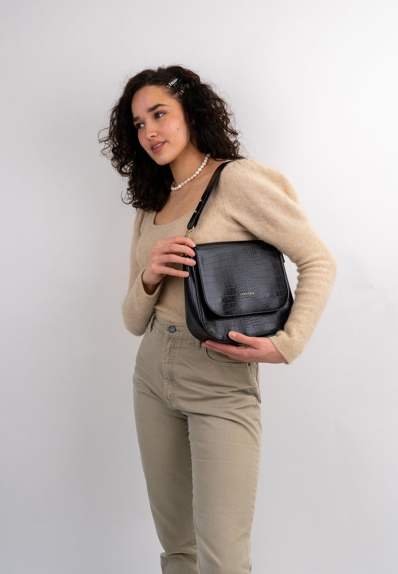 Black faux leather shoulder bag with a crocodile texture, featuring a front flap and gold hardware, held by a person wearing a beige top.