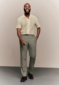 Beżowa koszula z krótkim rękawem o wzorzystym, fakturowanym designie, zestawiona z dopasowanymi jasnoszarymi spodniami i ciemnobrązowymi butami.