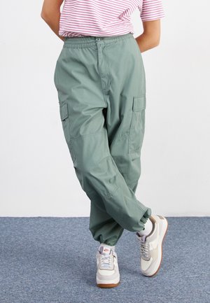Persona con pantalones cargo verdes, zapatillas blancas y beige, y una camisa rosa a rayas, de pie con las piernas cruzadas sobre una alfombra azul.