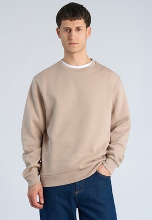 Junger Mann, der einen beigen Pullover und blaue Jeans trägt, steht mit einer Hand in der Tasche vor einem einfarbigen Hintergrund.