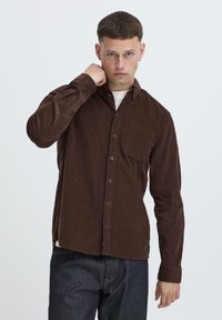 Brown corduroy shirt with a button-up front, single chest pocket, and long sleeves, featuring a textured fabric and slightly curved hem.